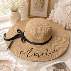 Könnte beinhalten: Ein beigefarbener Strohhut mit breiter Krempe und einem schwarzen Band, das an der Seite zu einer Schleife gebunden ist. Der Name Amelia ist in schwarzer Schreibschrift auf die Vorderseite der Krempe gestickt. Der Hut hat eine runde Krone.