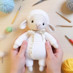May include: A white crocheted lamb stuffed animal with black bead eyes and a pink stitched nose. The lamb features floppy ears and a cream-colored ribbon with a small brown leaf pattern tied around its neck, crafted with a textured yarn finish.