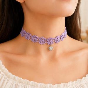 May include: A light purple lace choker necklace featuring a repeating floral pattern and a small, smooth silver-toned heart charm suspended from the center. The delicate crochet-style fabric sits snugly around the neck, showcasing intricate detail.