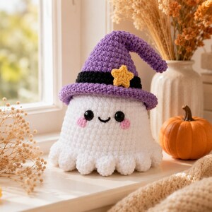 Puede incluir: Un juguete de peluche de fantasma tejido a ganchillo en color blanco con un sombrero de bruja morado, una banda negra y una peque&ntilde;a estrella amarilla. El fantasma tiene dos ojos negros circulares, una sonrisa negra y mejillas rosadas.