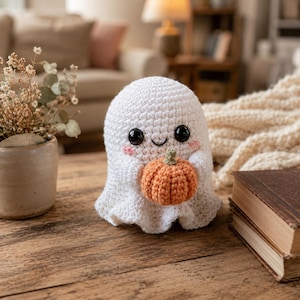 Op de afbeelding: Een wit gehaakt spookfiguurtje met grote zwarte ogen en roze wangen, dat een kleine oranje gehaakte pompoen vasthoudt. Het spookje heeft een ronde kop en een gedrapeerd lichaam, gemaakt met zichtbare steken op een houten ondergrond.