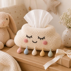 May include: A cream-colored crocheted tissue box cover shaped like a cloud with a smiling face, closed eyes, and pink cheeks. Five small, multi-colored crocheted raindrops in pink, blue, cream, yellow, and green hang from the bottom edge.