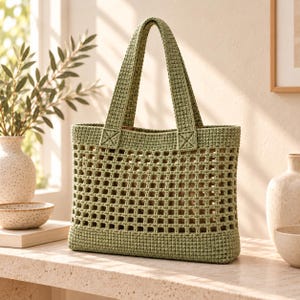 May include: A sage green crochet tote bag featuring a structured rectangular base, a dense stitch pattern at the bottom and top edges, and a wide open-work grid design in the center. The bag includes two sturdy matching crochet handles.