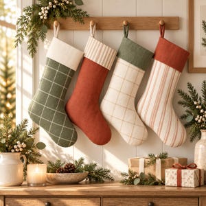 May include: Four hanging Christmas stockings in a row on a wooden rack. The first is green with a white grid and cream cuff, the second is solid rust with a striped cuff, the third is cream with a green grid and green cuff, and the fourth is striped with a rust cuff.