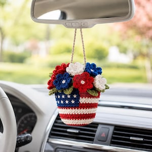 May include: A crocheted hanging ornament featuring a small pot designed with the American flag pattern, filled with a bouquet of red, white, and blue crocheted flowers and green leaves, suspended by a tan cord from a vehicle rearview mirror.