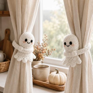 May include: Two white crocheted ghost curtain tiebacks featuring black embroidered eyes, a small smile, and pink blush cheeks. Each ghost has long, wrap-around arms that secure the light beige fabric curtains in place on either side of a window.