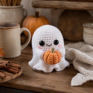 Puede incluir: Una figura de fantasma tejida a ganchillo en color blanco con grandes ojos de seguridad negros, una peque&ntilde;a sonrisa bordada y detalles rosados en las mejillas. El fantasma sostiene una peque&ntilde;a calabaza tejida naranja con un tallo verde.