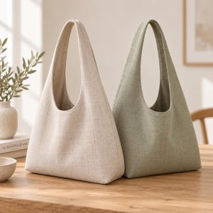 May include: Two hobo-style shoulder bags made of a textured fabric. One bag is a light beige color, and the other is a muted sage green. Both bags feature a single integrated handle, a wide opening, and a structured base with a clean, simple design.