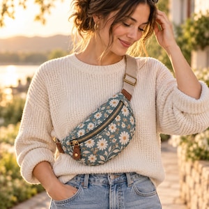 Peut inclure: Un sac banane bleu sarcelle orné d'un motif floral de marguerites blanches, avec des détails en cuir marron et une sangle beige réglable. Le sac possède une fermeture éclair dorée et est porté en bandoulière sur un pull en maille crème et un jean.