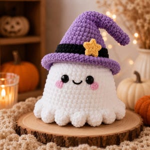 Puede incluir: Un juguete de peluche de fantasma tejido a ganchillo en color blanco, con un sombrero puntiagudo morado, una banda negra y una peque&ntilde;a estrella amarilla. El fantasma tiene ojos negros circulares, una sonrisa bordada y mejillas rosadas.