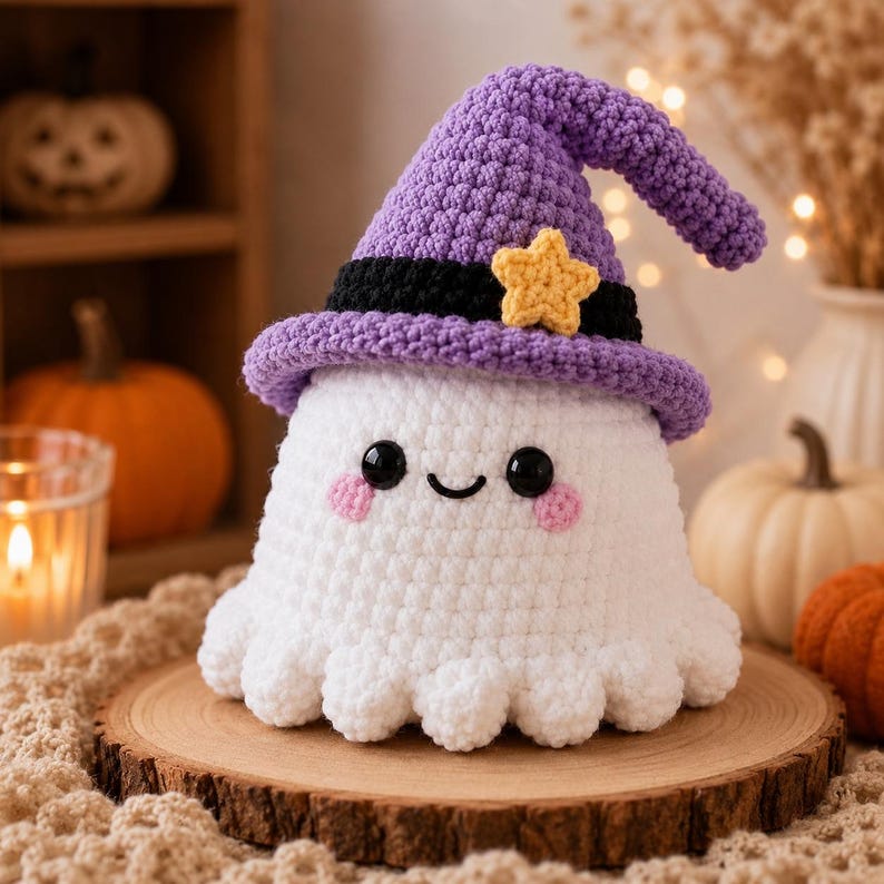 Puede incluir: Un juguete de peluche de fantasma tejido a ganchillo en color blanco, con un sombrero puntiagudo morado, una banda negra y una peque&ntilde;a estrella amarilla. El fantasma tiene ojos negros circulares, una sonrisa bordada y mejillas rosadas.