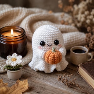 Puede incluir: Una figura de fantasma tejida a ganchillo en color blanco con ojos de cuentas negras y mejillas rosadas, sosteniendo una peque&ntilde;a calabaza tejida en color naranja. El fantasma se sit&uacute;a junto a una vela encendida en un frasco de vidrio marr&oacute;n.