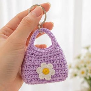 Può includere: Un piccolo portachiavi a forma di borsa lavorato all'uncinetto in color viola chiaro, con un'applicazione a margherita bianca con centro giallo. La borsa in miniatura ha un manico arrotondato ed è fissata a un anello metallico argentato.