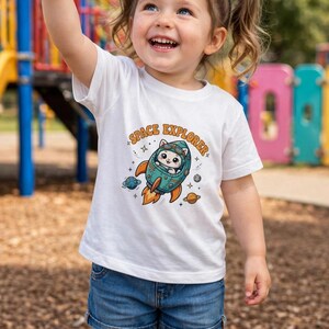 Peut inclure: Un t-shirt blanc à manches courtes avec un motif de chat dans une fusée bleu sarcelle et orange portant l'inscription SPACE EXPLORER, entouré de petites étoiles et de planètes, porté avec un short en jean bleu et des nœuds orange.