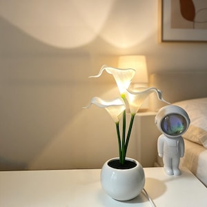 Puede incluir: Una maceta esférica blanca que contiene tres flores de cala artificiales iluminadas con tallos verdes. Junto a la maceta hay una pequeña figura de astronauta blanca con una visera reflectante e iridiscente en el casco.