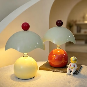 May include: Two mushroom-shaped table lamps with pale green scalloped shades and spherical finials. The left lamp has a yellow base, while the right lamp has an orange base and sits on a book next to a small white and gold astronaut figurine.