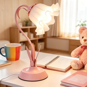 May include: A pink table lamp shaped like a flower with a curved stem and a ruffled, translucent white glass shade that emits a soft glow. The base is circular and pink, featuring decorative leaf-like accents at the bottom of the stem.