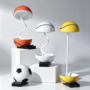 Puede incluir: Tres lámparas de mesa con forma de balones de baloncesto en naranja, blanco y amarillo, cada una con base negra. Dos lámparas tienen tallos blancos flexibles, una tiene un tallo recto. Un balón de fútbol blanco y negro sobre base negra.