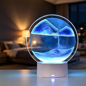 May include: A circular glass sand art lamp featuring blue and white sand forming mountain and water patterns. The round frame is illuminated by a bright white LED light, and the entire structure is supported by a solid white cylindrical base.