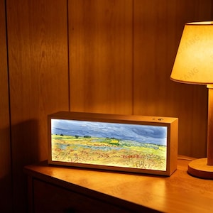 Pode incluir: Uma caixa de luz retangular com moldura de madeira exibindo uma pintura de paisagem de um campo sob um céu tempestuoso. A iluminação interna destaca os tons azuis, verdes e amarelos da obra. Um pequeno botão é visível na borda superior.