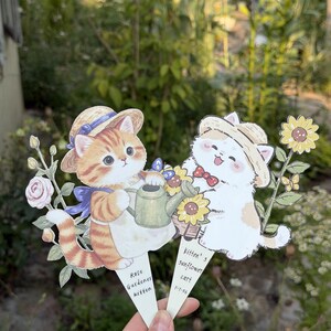 May include: Two die-cut paper garden markers featuring illustrations of kittens in straw hats. One orange tabby kitten holds a green watering can with the text 'Rose Gardener kitten'. A white kitten stands by a sunflower cart with the text 'kitten's sunflower cart'.