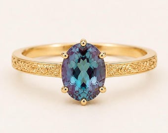 May include: A gold ring featuring an oval-cut, color-changing gemstone that shifts between shades of teal, blue, and purple. The band is crafted from yellow gold and displays an intricate, engraved scrollwork pattern along the entire shank.