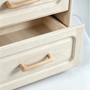 Peut inclure: Une commode en bois clair dot&eacute;e de deux tiroirs aux bords arrondis et lisses avec un design &agrave; panneau encastr&eacute;. Chaque tiroir est &eacute;quip&eacute; d'une poign&eacute;e incurv&eacute;e en bois naturel qui s'harmonise avec le grain clair des fa&ccedil;ades.