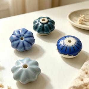 Peut inclure: Quatre boutons en céramique avec un trou central, chacun présentant une finition dans les tons bleus. Un est bleu clair, un bleu moyen avec des nuances radiales, un bleu sarcelle foncé avec un vernis moucheté, et un à base blanche avec des motifs de pétales bleus.