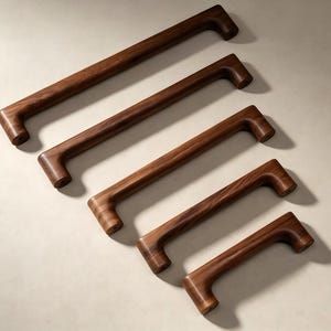 Peut inclure: Cinq poignées de placard en bois brun foncé disposées en diagonale, de la plus longue à la plus courte. Chaque poignée présente un design en barre rectangulaire lisse avec des bords arrondis et des points de fixation cylindriques aux extrémités.