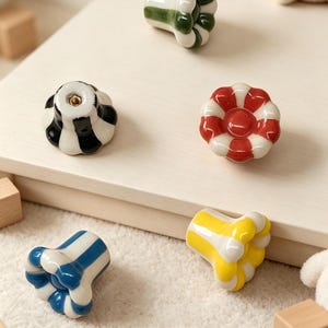 Peut inclure: Un ensemble de cinq boutons de tiroir en céramique, chacun présentant une forme festonnée en fleur avec des rayures verticales alternant les couleurs vert foncé, noir, rouge, bleu et jaune sur une base blanche brillante avec fixation.