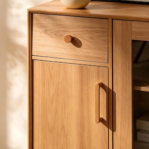 Peut inclure: Un buffet en bois de ch&ecirc;ne clair comprenant un tiroir sup&eacute;rieur avec un bouton rond en bois et une porte de placard en dessous munie d'une poign&eacute;e verticale en bois. Le meuble pr&eacute;sente une section vitr&eacute;e sur la droite avec des &eacute;tag&egrave;res.