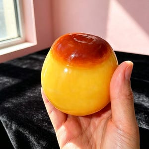 Puede incluir: Un objeto liso y redondeado con un acabado brillante, que presenta una base de color amarillo vibrante que se transforma en un tono ámbar-marrón profundo y translúcido en la parte superior. La superficie es pulida y refleja tonos cálidos.