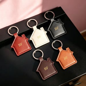 May include: Five house-shaped keychains in red, cream, black, dark brown, and tan colors. Each keychain features a silver metal ring and the letters BF embossed in gold on the front. The keychains are made of a smooth material with visible stitching.