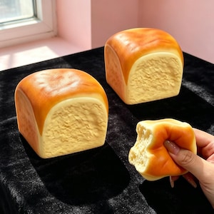 May include: Two square-shaped, golden-brown bread loaves with textured, pale yellow centers. A smaller, squishy piece of the same bread is held, showing a porous, light-colored interior texture. The items have a realistic, baked appearance.