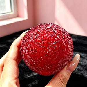 May include: A spherical object with a deep red, translucent, and highly textured surface. The exterior features numerous small, jagged, and crystalline protrusions that catch the light, creating a multifaceted appearance across the entire round form.