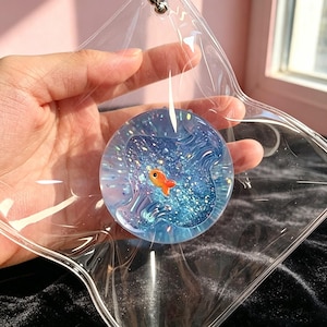 May include: A circular, transparent acrylic charm featuring a blue, glitter-filled liquid center with a small orange goldfish figurine inside. The charm is encased in a clear, star-shaped plastic pouch with a metal ring attachment at the top.