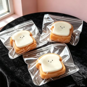May include: Three individual pieces of soap shaped like slices of toast, each packaged in a clear plastic zip-top bag. The soap features a golden-brown crust and a smooth, cream-colored center with a simple, printed black smiley face design.