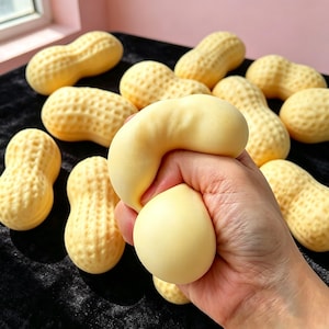 Può includere: Una collezione di giocattoli antistress a forma di arachide, di colore giallo pallido, con una superficie ruvida simile a un guscio. Uno dei giocattoli morbidi e comprimibili viene schiacciato, mostrandone il materiale flessibile e deformabile.
