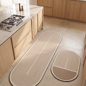 Op de afbeelding: Een set van twee vloermatten met een beige en crèmekleurig palet. De matten hebben een herhalend patroon van gebogen lijnen met een donkere rand. De ene mat is lang en ovaal, terwijl de andere kleiner en ovaal is met een effen beige gedeelte.