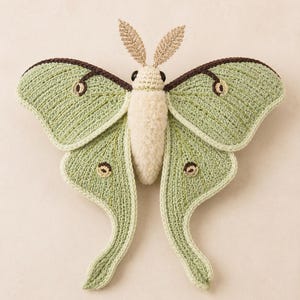 Pode incluir: Uma mariposa-lua de crochê com asas verde-claras, bordas castanho-escuras e manchas oculares circulares. O corpo é feito de fio texturizado creme, as antenas são de fio dourado metálico e os olhos são contas pretas.