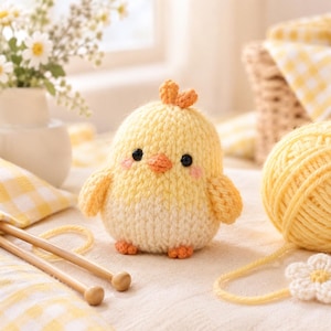 May include: A small, round, hand-knitted chick figurine featuring a soft yellow and cream-coloured yarn body, two black bead eyes, a small orange beak, tiny orange feet, and a small tuft of orange yarn on top of its head with pink blush accents.