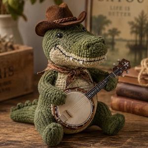 Può includere: Un giocattolo a forma di alligatore verde all'uncinetto che indossa un cappello da cowboy in feltro marrone e un fazzoletto da collo fantasia. L'alligatore ha la pancia color crema e tiene in mano un piccolo banjo in legno.