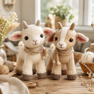 Könnte beinhalten: Zwei gestrickte Ziegen-Stofftiere stehen nebeneinander. Die linke Ziege ist cremefarben mit einem passenden Schal, während die rechte Ziege hellbraun mit einem weißen Streifen im Gesicht und einem passenden Schal ist. Beide haben kleine Hörner und rosa Ohren.