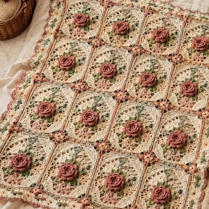May include: A square crochet blanket featuring a grid of cream-colored panels, each adorned with a three-dimensional dusty pink rose and green leaf embroidery. The edges are finished with a delicate scalloped border in pink and cream yarn.