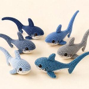 May include: Six small, handmade crochet shark toys in various shades of blue and gray. Each shark features a rounded body, a dorsal fin, a long tail, and a white underbelly with black plastic safety eyes and stitched mouth details.