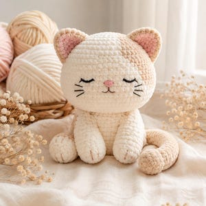 Pode incluir: Um peluche de gato feito em croché com fio creme e castanho claro, com orelhas internas cor-de-rosa, nariz cor-de-rosa e olhos e bigodes pretos bordados. O gato está sentado numa pose neutra com a cauda enrolada, feito de fio macio.