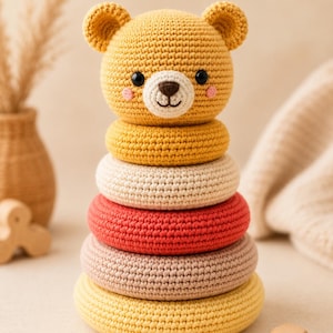 May include: A crochet stacking toy featuring a yellow bear head topper with black eyes and a brown nose. The toy consists of four stacked rings in yellow, cream, red, and light brown, all crafted with a textured yarn finish and a rounded shape.
