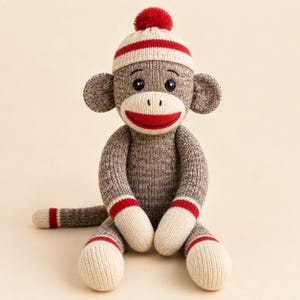 Puede incluir: Un peluche de mono hecho de tejido de punto marrón, crema y rojo. El mono presenta una cara de color crema, una boca roja ancha, ojos de botón negros y un gorro de punto a juego con un pompón rojo en la parte superior. Tiene brazos y piernas largos.