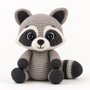 Puede incluir: Un peluche de mapache tejido a ganchillo con cuerpo gris claro, patas gris oscuro y una cola a rayas. La cara tiene un hocico blanco, parches negros alrededor de los ojos y grandes ojos de plástico negro. Orejas puntiagudas y nariz bordada.