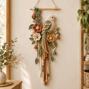 Puede incluir: Un tapiz de macramé que presenta un pájaro de color crema posado en una rama, rodeado de flores de ganchillo en tonos terracota, amarillo mostaza y blanco, con hojas verde salvia y cordones colgantes sujetos a un soporte de madera.